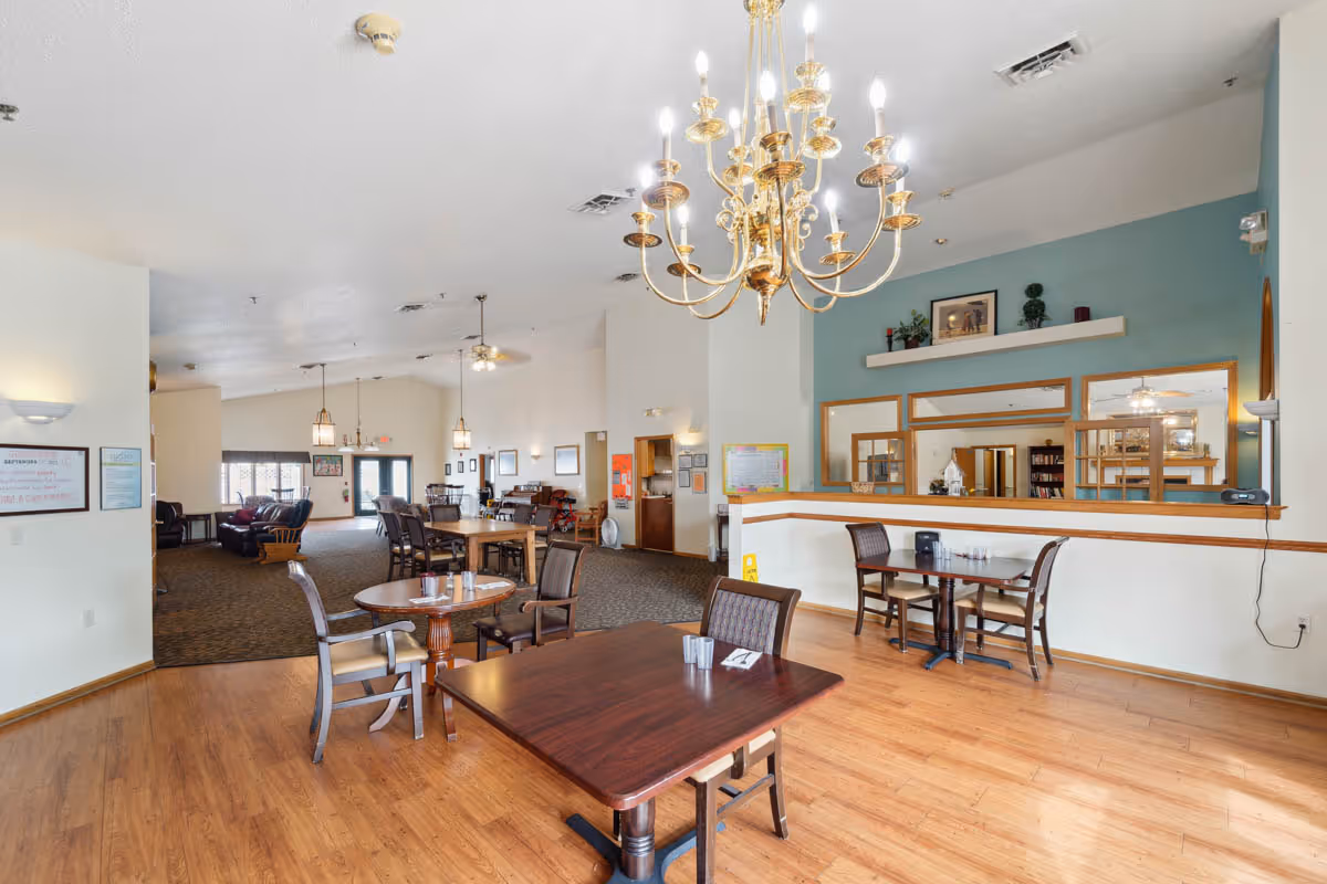A spacious common area in a senior living facility featuring wooden floors and carpeted seating areas. There are several tables with chairs arranged for dining or socializing. A large gold chandelier hangs from the ceiling, and the walls are decorated with framed pictures and plants. The room has a welcoming atmosphere with a mix of natural and artificial lighting.