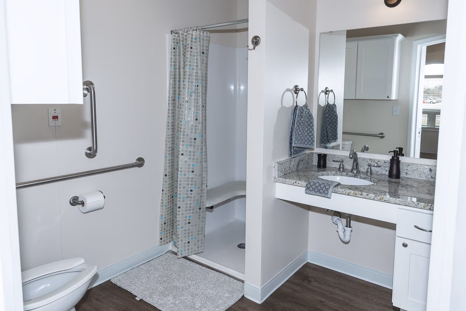 A clean and accessible bathroom featuring a toilet with grab bars, a walk-in shower with a bench and a patterned shower curtain, a granite countertop with a sink, hand soap dispenser, and matching hand towels, and a large mirror above the sink. The floor is wood-style and there is a white rug in front of the shower.