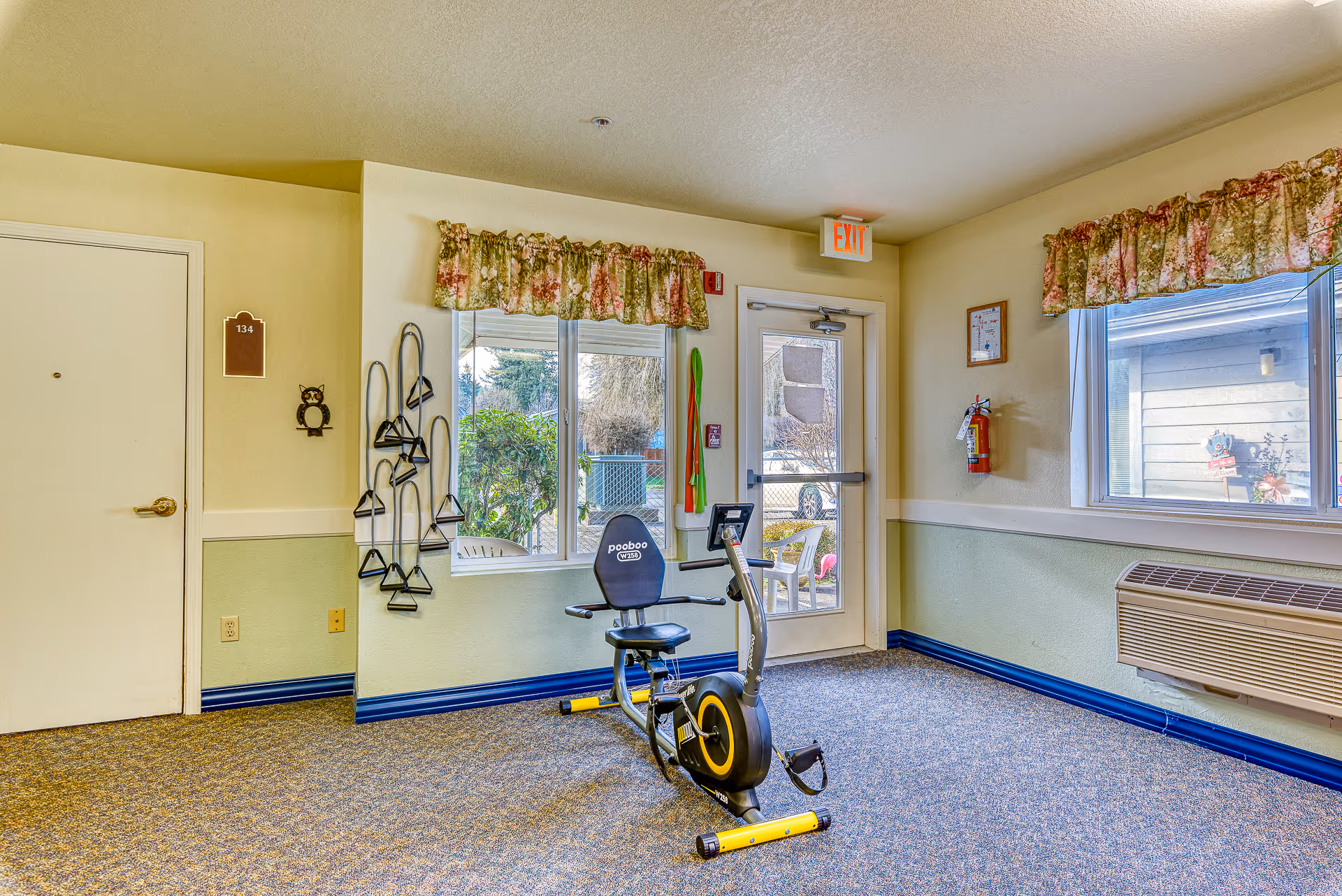 A small exercise room in a senior living facility with a recumbent exercise bike positioned in the center. The room has beige walls with a green lower half, floral valance curtains on the windows, and a door with an exit sign above it. Exercise bands hang on the wall next to a window, and there is a fire extinguisher mounted on the wall. Outside the windows, some outdoor furniture and greenery are visible.