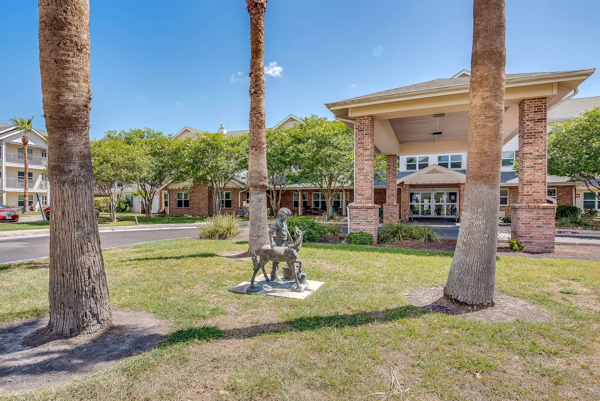 Exterior view of Solstice Senior Living at Corpus Christi showing the entrance with a covered drop-off area supported by brick columns. In front of the entrance is a grassy area with a bronze statue of a child and a dog, surrounded by palm trees and other greenery under a clear blue sky.