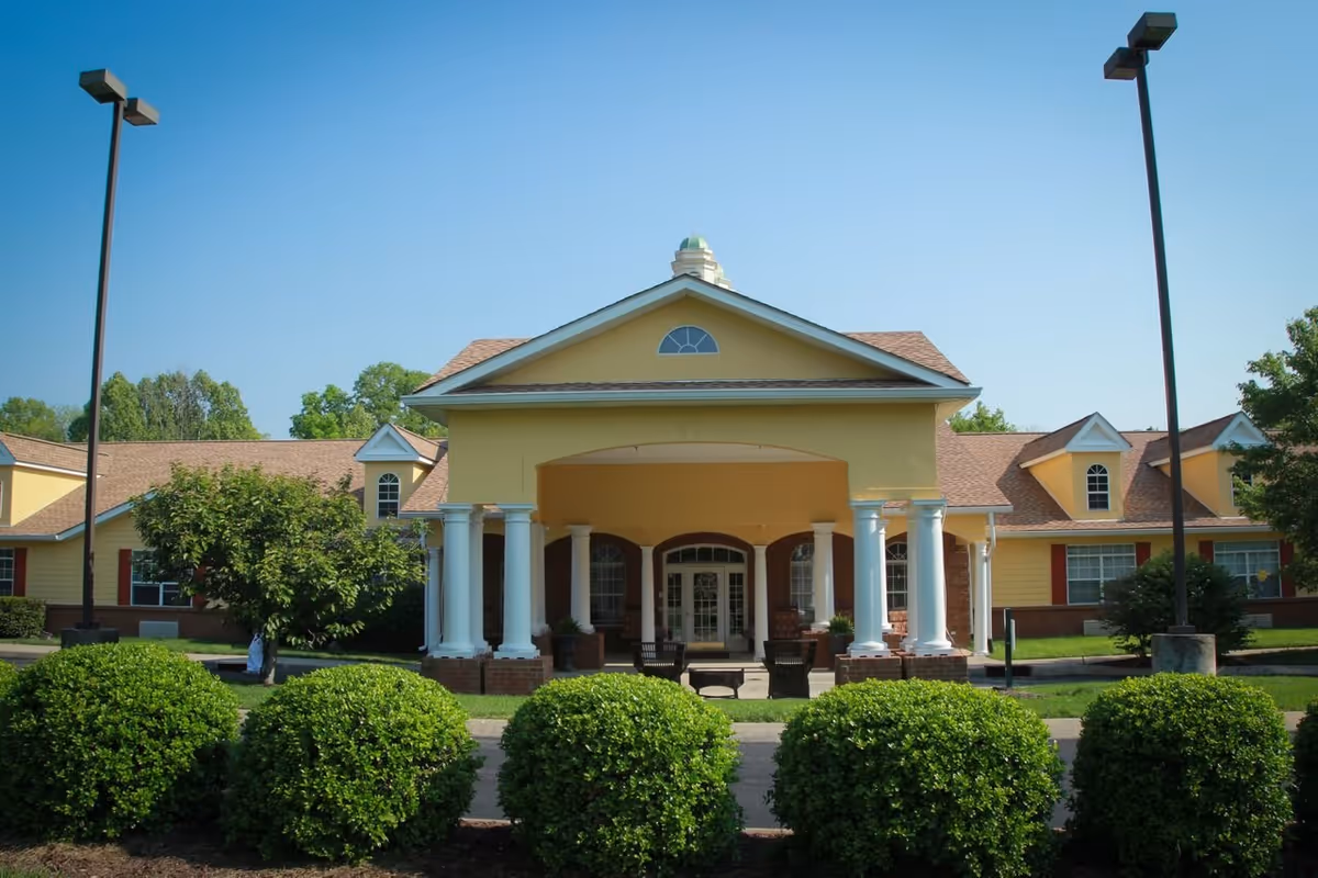 Front entrance of a yellow senior living building with white columns, a covered portico, and trimmed shrubs.