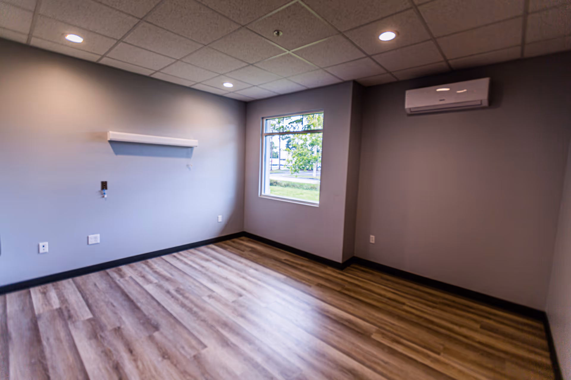 Empty room with gray walls, wood-patterned flooring, a window showing greenery outside, recessed ceiling lights, and a wall-mounted air conditioning unit.