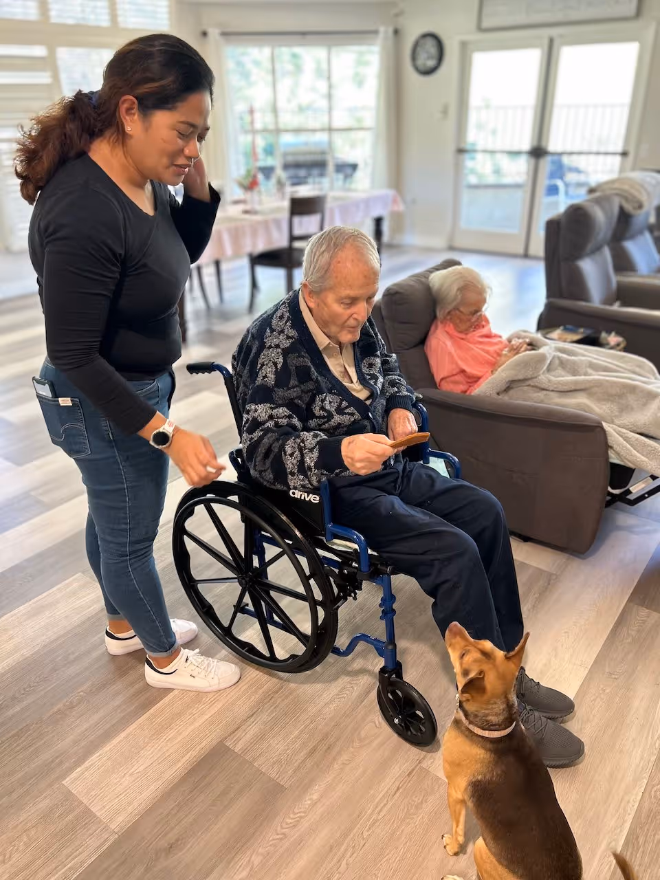 An elderly man in a wheelchair is interacting with a small dog while a woman stands nearby. In the background, an elderly woman is sitting in a recliner chair covered with a blanket. The setting appears to be a bright, spacious living room with wooden flooring and large windows.