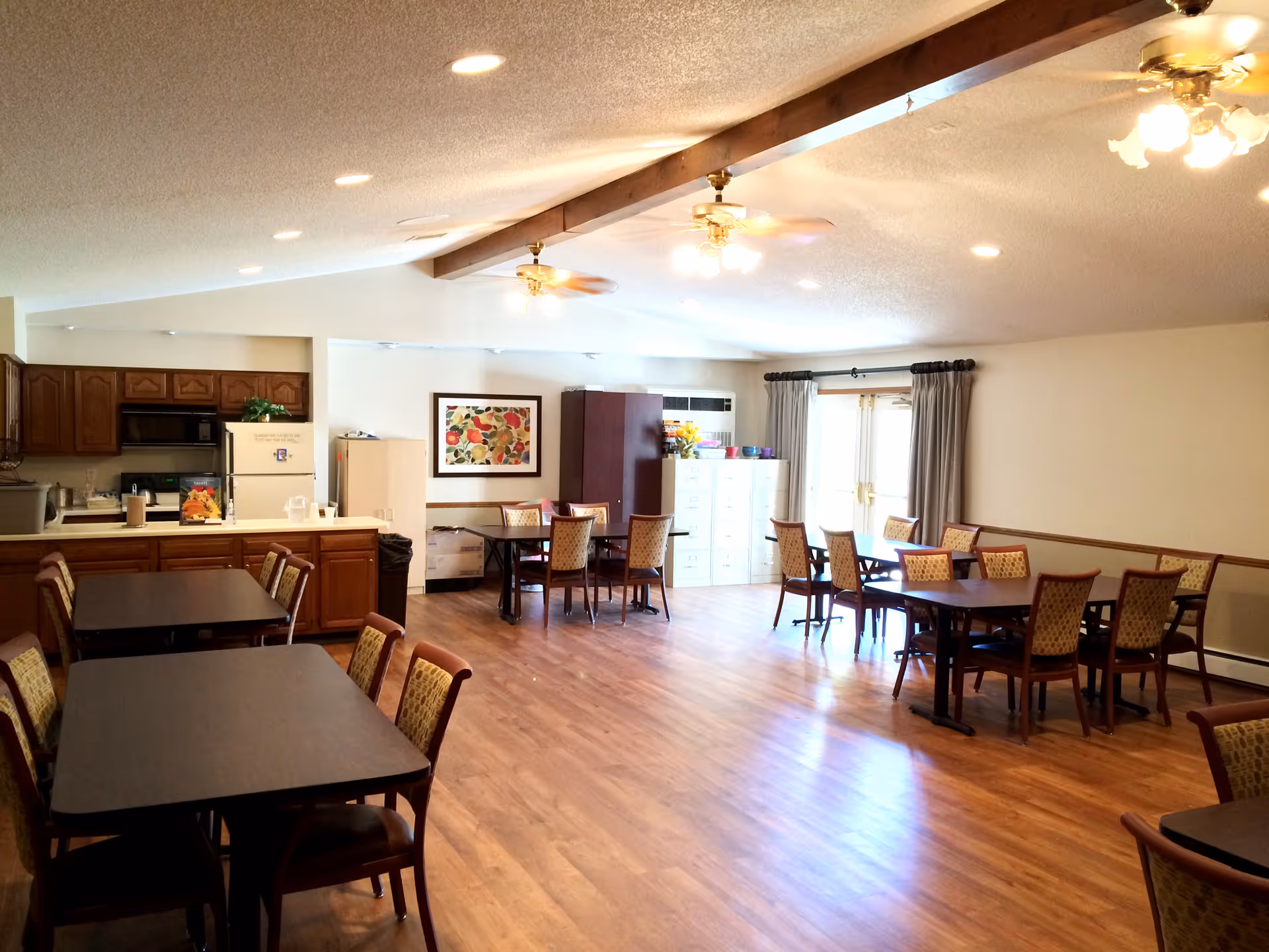 A spacious dining room with several dark wood tables and patterned chairs arranged neatly. The room has wooden flooring, ceiling fans with lights, and large windows with curtains letting in natural light. A kitchen area with wooden cabinets, a refrigerator, and a microwave is visible on the left side. There is a colorful framed artwork on the wall and filing cabinets near the windows.