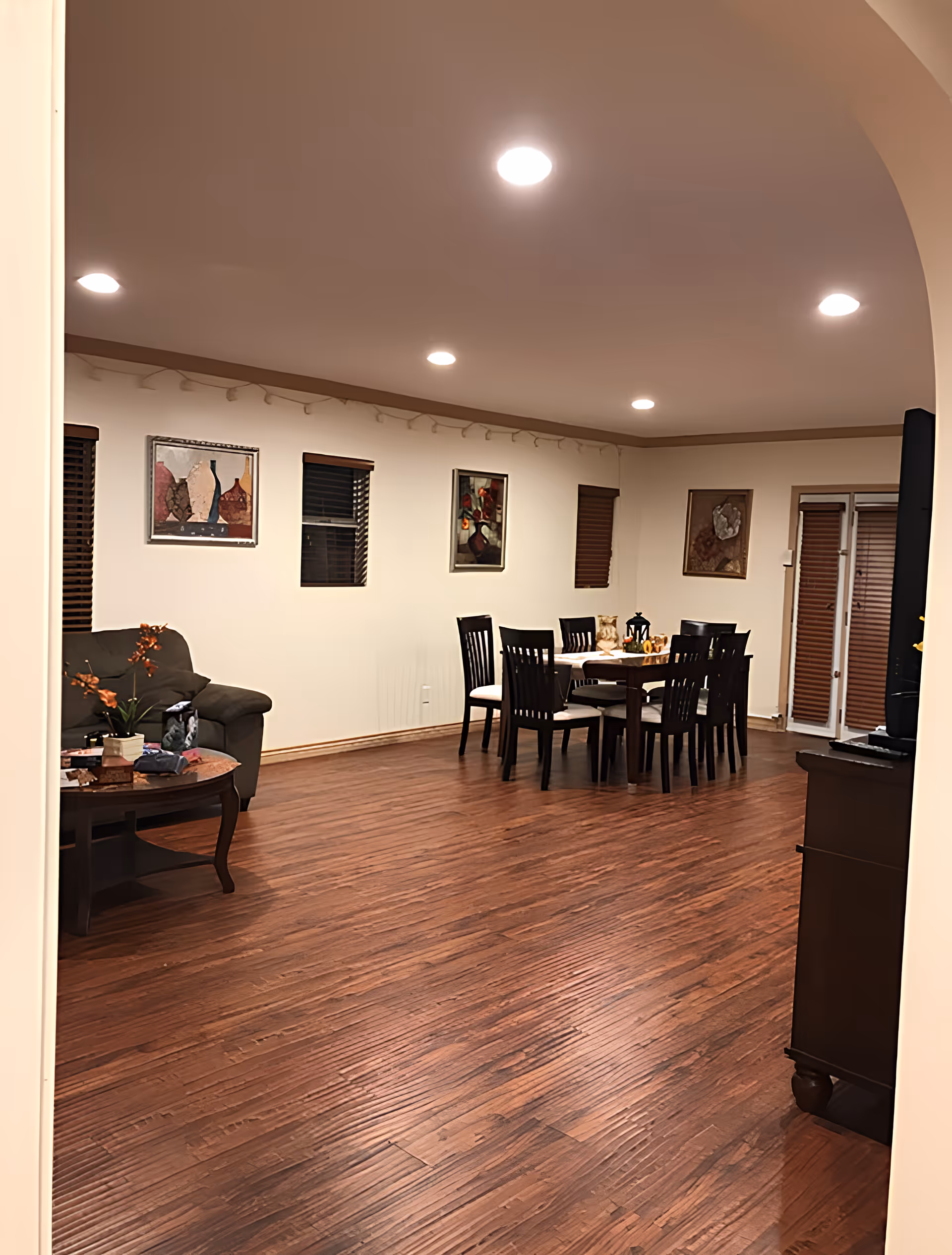 Interior view of a senior living facility named Paradise showing a spacious room with wooden flooring, a dining table with eight chairs, a green armchair, a coffee table with a plant and books, several framed artworks on the walls, and recessed ceiling lights.