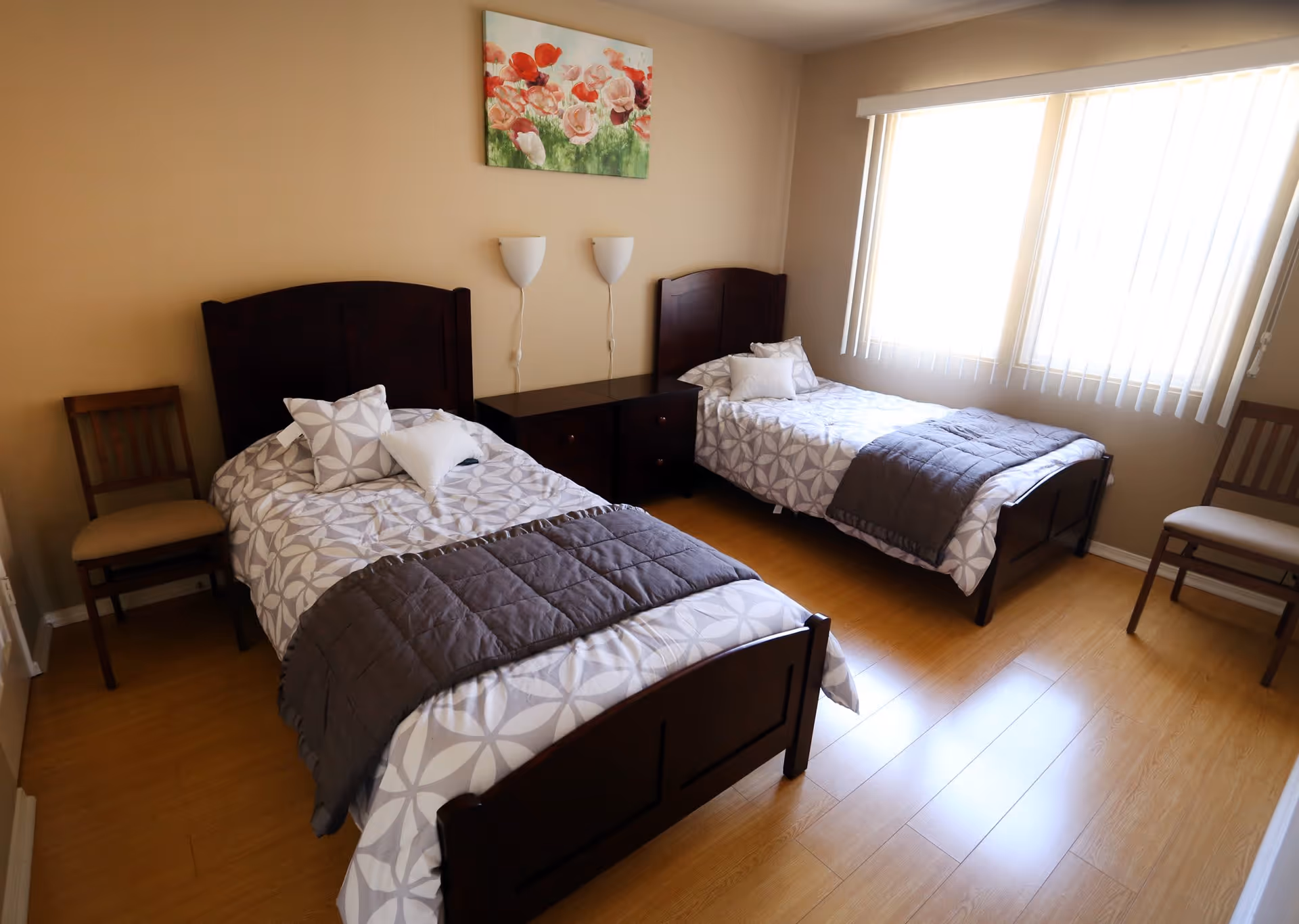 A bedroom with two single beds featuring dark wooden headboards and footboards, each bed made with patterned white and gray bedding and gray blankets. Between the beds is a dark wooden nightstand with two drawers. Two wall-mounted white lamps are above the nightstand. There are two wooden chairs with beige cushions, one on each side of the room. A large window with vertical blinds allows natural light to fill the room. A floral painting with red and pink flowers hangs on the beige wall above the nightstand.