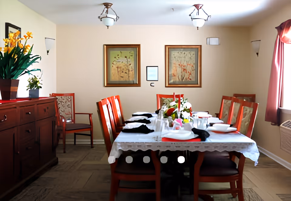 Dining room with a table set for a meal, red wooden chairs, a sideboard with plants, and framed artwork on the wall.