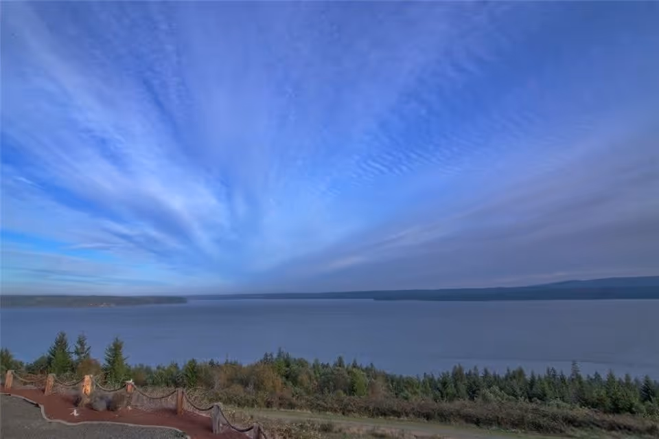 A scenic view overlooking a large body of water with a forested shoreline in the foreground and a partly cloudy blue sky above.