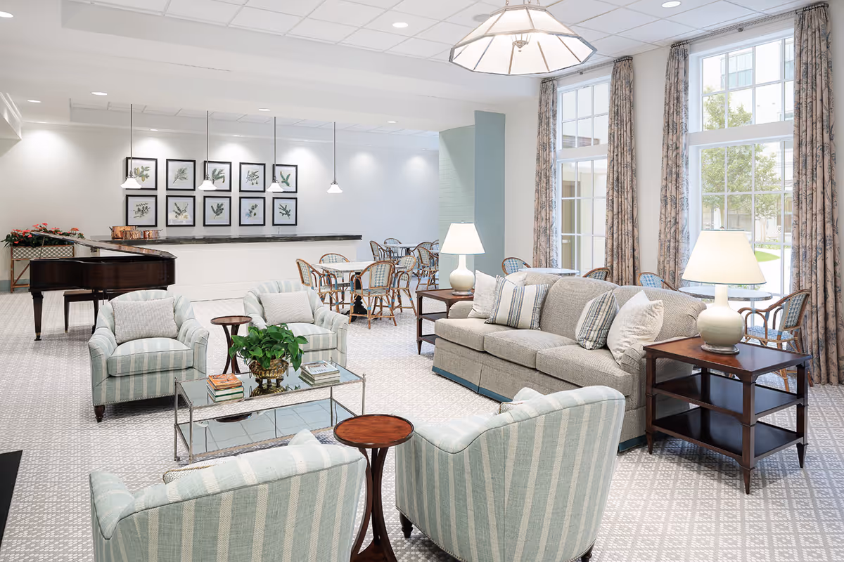 A bright and spacious living room area in a senior living facility featuring a beige sofa with striped and solid pillows, four striped armchairs arranged around a glass coffee table with books and a plant, two wooden side tables with lamps, large windows with patterned curtains letting in natural light, a grand piano, and a dining area with tables and chairs in the background.