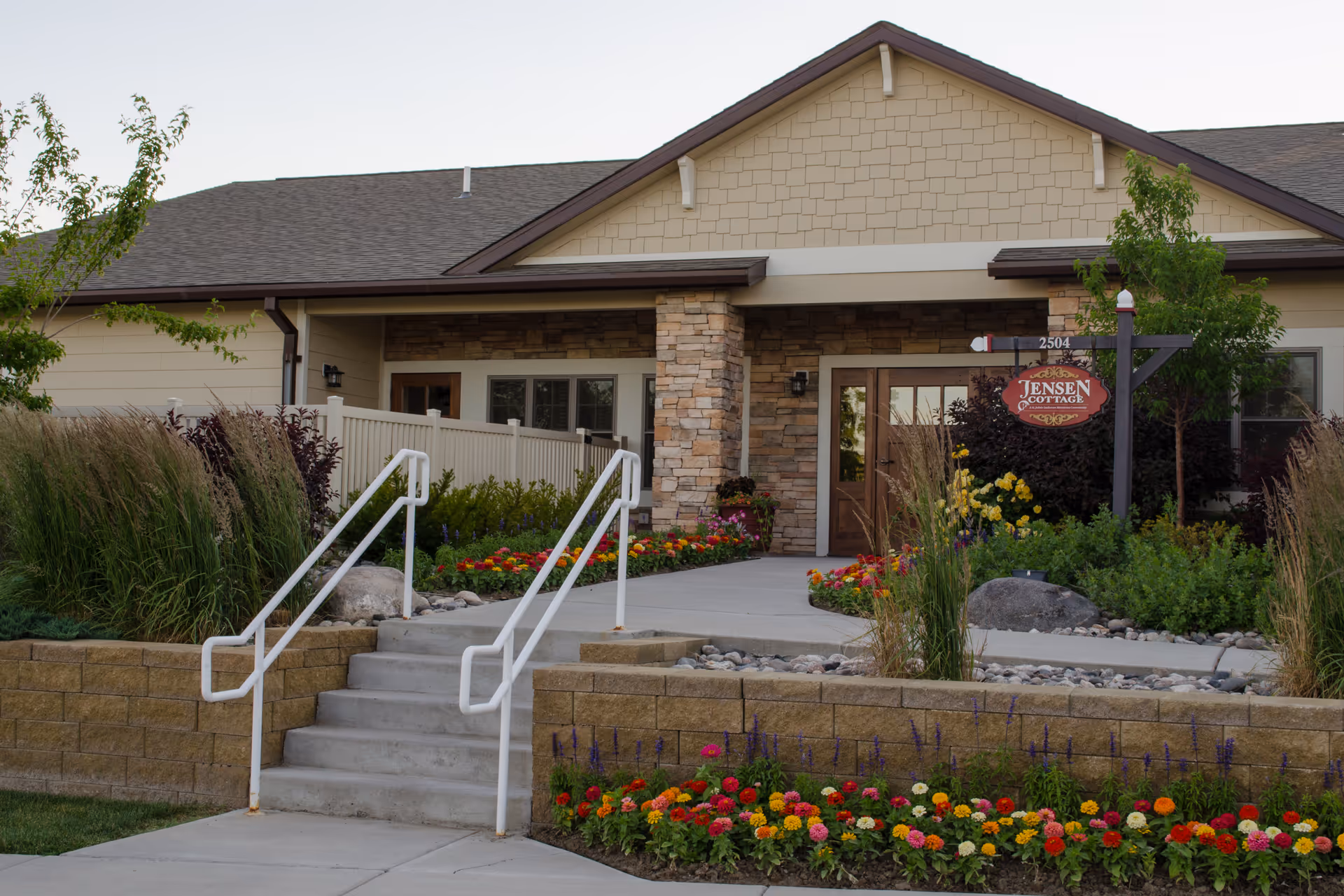 Entrance to a single-story building with stone and beige siding, featuring a small staircase with white railings leading up to a walkway surrounded by colorful flowers and greenery. A sign near the door reads 'Jensen Cottage'.