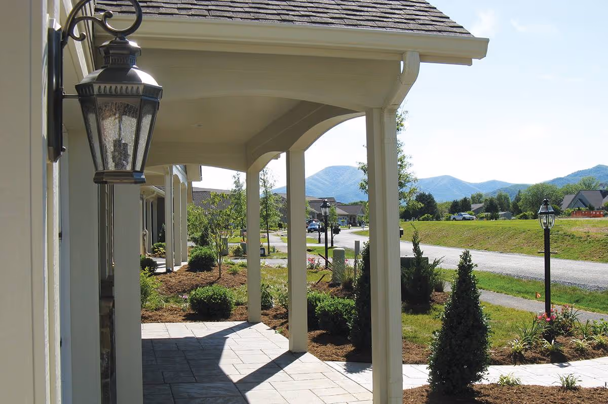 Covered walkway with columns and outdoor lanterns at the entrance of a building, landscaped garden beds with shrubs and small trees, a paved road, and mountains in the background under a clear sky.