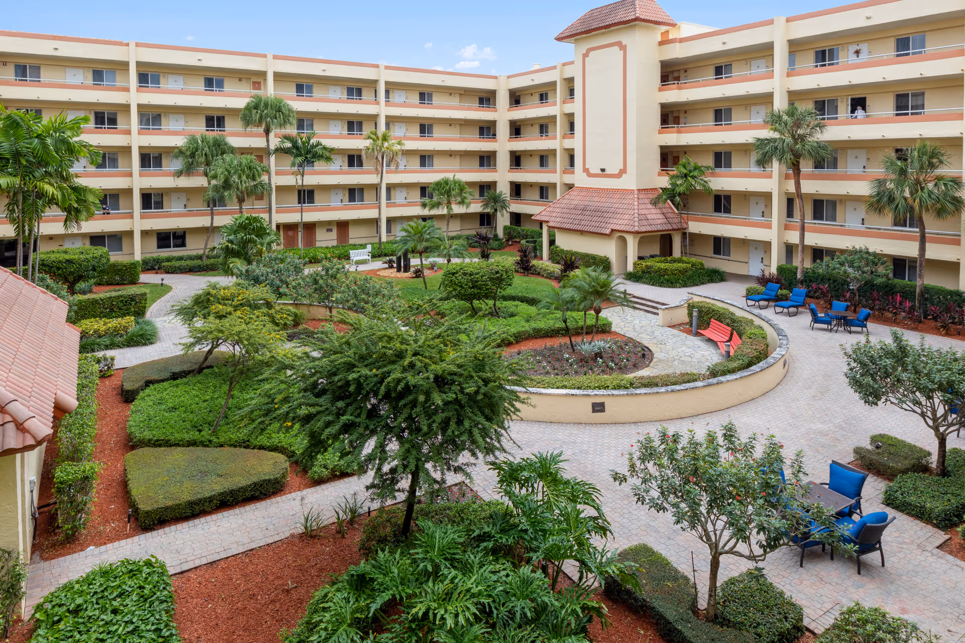 View of a landscaped courtyard in a multi-story residential building with pathways, green bushes, palm trees, benches, and outdoor seating areas with blue and red chairs under a clear blue sky.