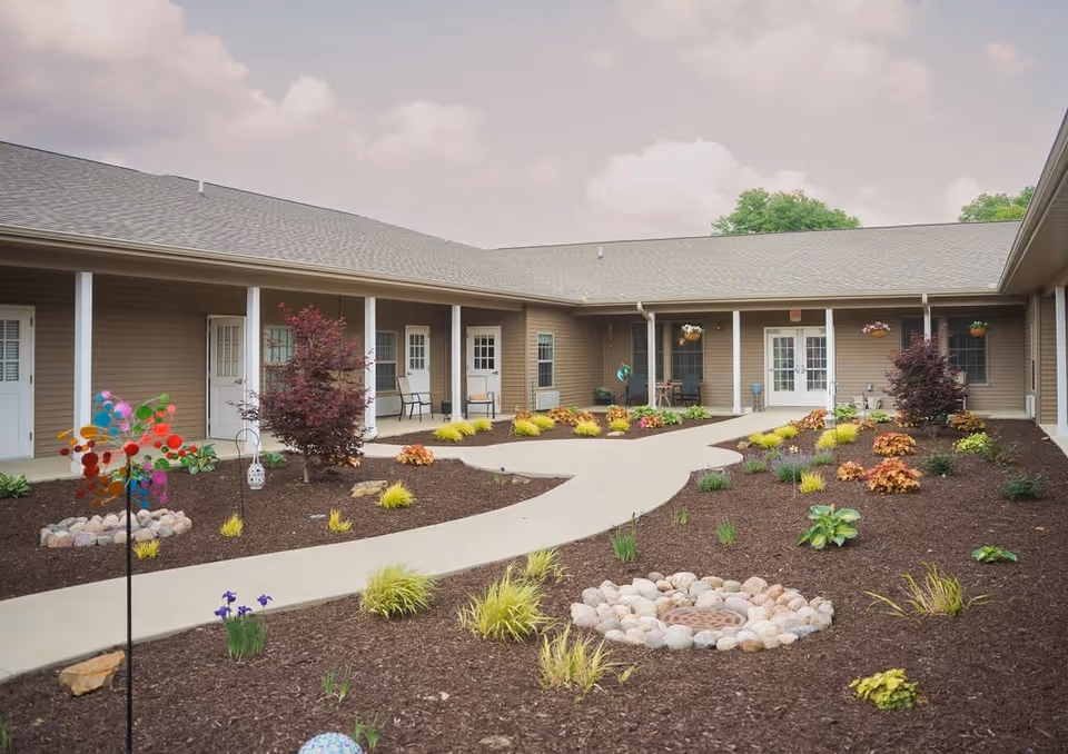 Outdoor courtyard area of a senior living facility with a paved walkway, landscaped garden beds with small plants and shrubs, and a building with multiple doors and windows surrounding the courtyard. There are chairs placed near some doors and hanging flower baskets on the building.