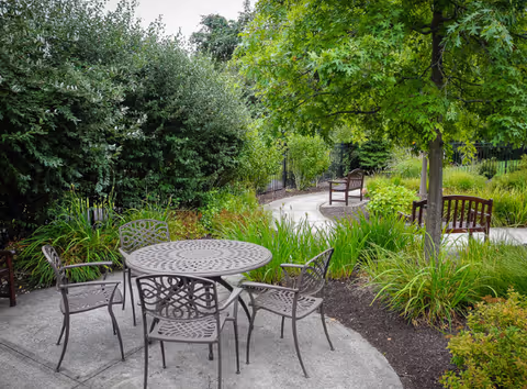 Outdoor patio area with a round metal table and four matching chairs on a concrete surface surrounded by green bushes, plants, and trees with additional wooden benches along a curved pathway.