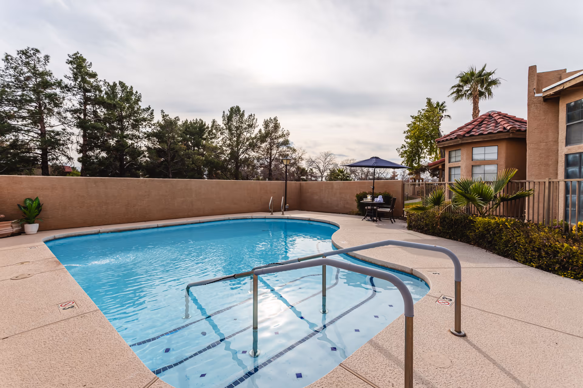 Outdoor swimming pool with metal handrails leading into the water, surrounded by a concrete deck. There are trees and bushes around the pool area, a small table with chairs and an umbrella near the back wall, and a building with a red-tiled roof and large windows on the right side.