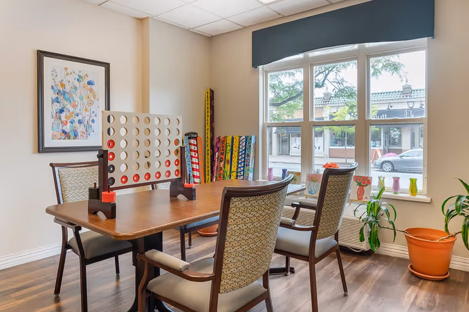A bright room with a wooden table and four upholstered chairs. On the table is a large Connect Four game set up with red and black discs. Behind the table is a window with a view of a street and buildings outside. The room has light-colored walls, a framed floral artwork, several colorful tall board games leaning against the wall, and potted plants near the window.