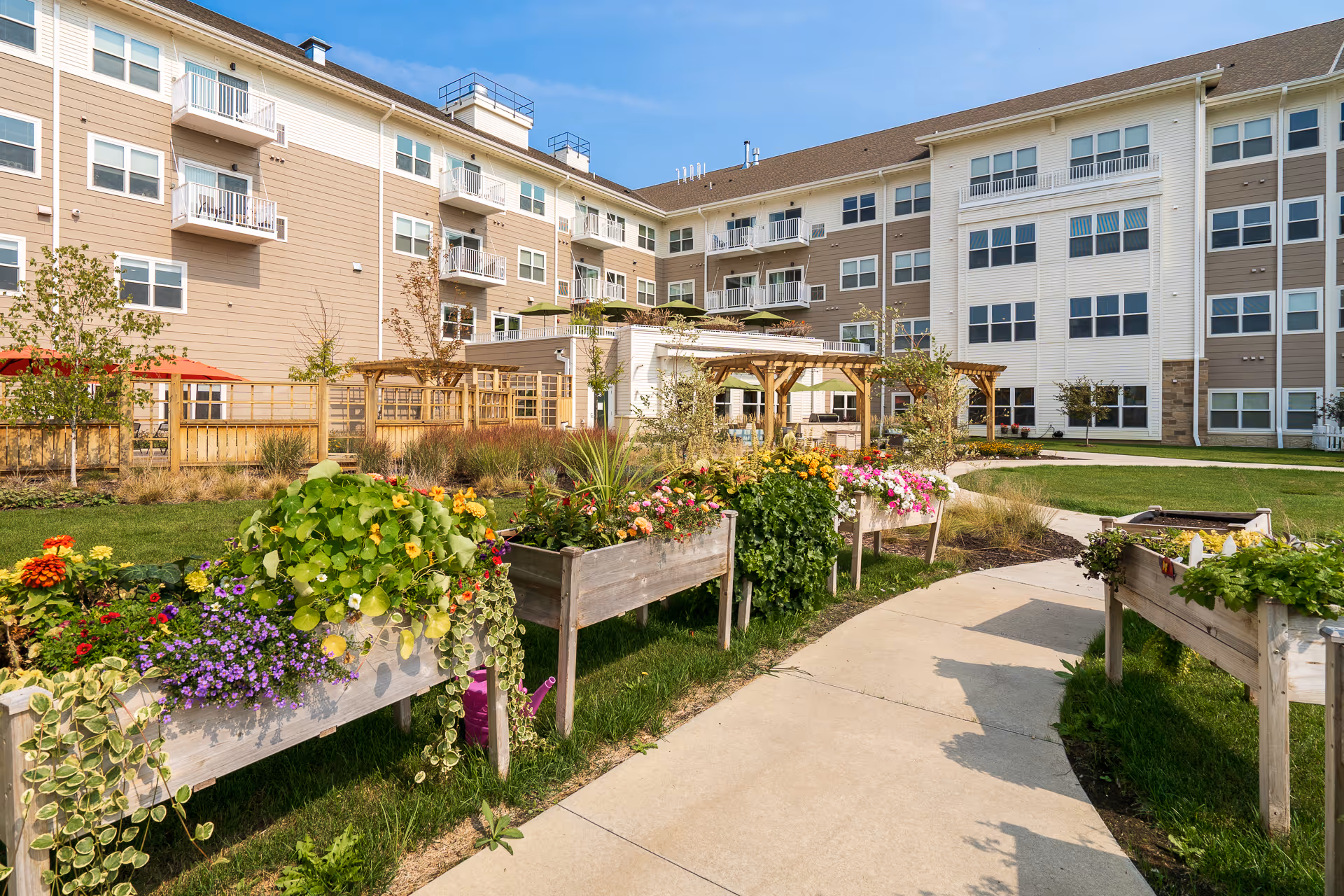 Outdoor garden area at Riley Crossing Senior Living with raised flower beds filled with colorful flowers and greenery, a paved walkway, and a multi-story residential building in the background under a clear blue sky.
