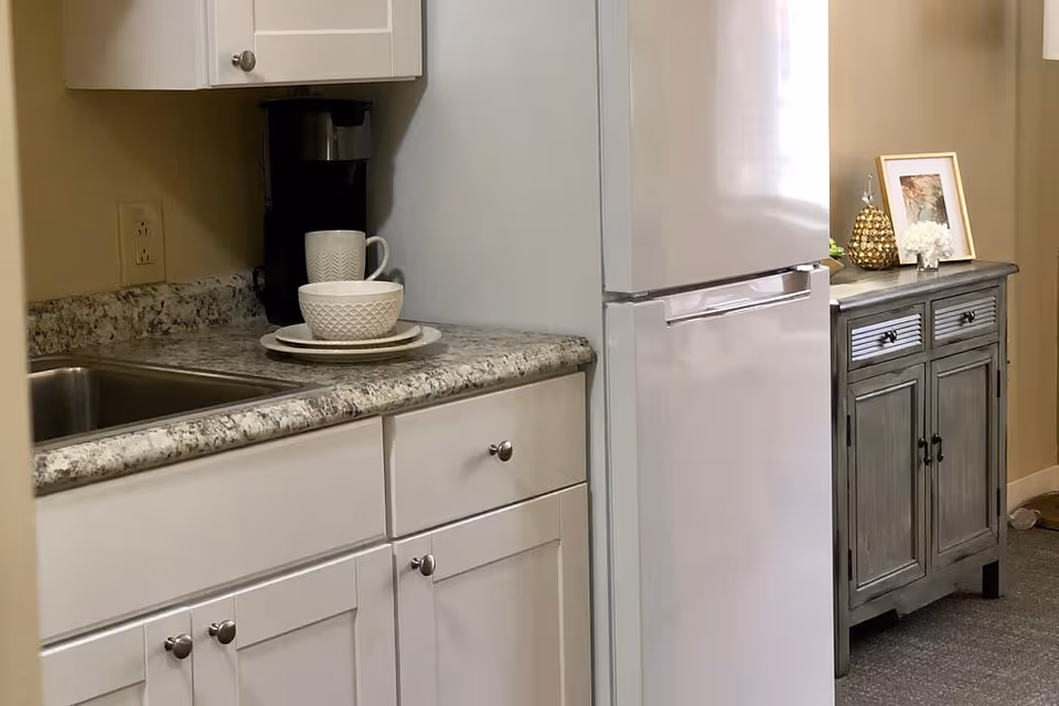 A small kitchen area with white cabinets, a granite countertop, a stainless steel sink, a coffee maker, and white dishware including a bowl, plate, and mug. Next to the kitchen is a white refrigerator and a gray wooden sideboard with decorative items including a framed picture, a golden pineapple, and a small white flower arrangement.
