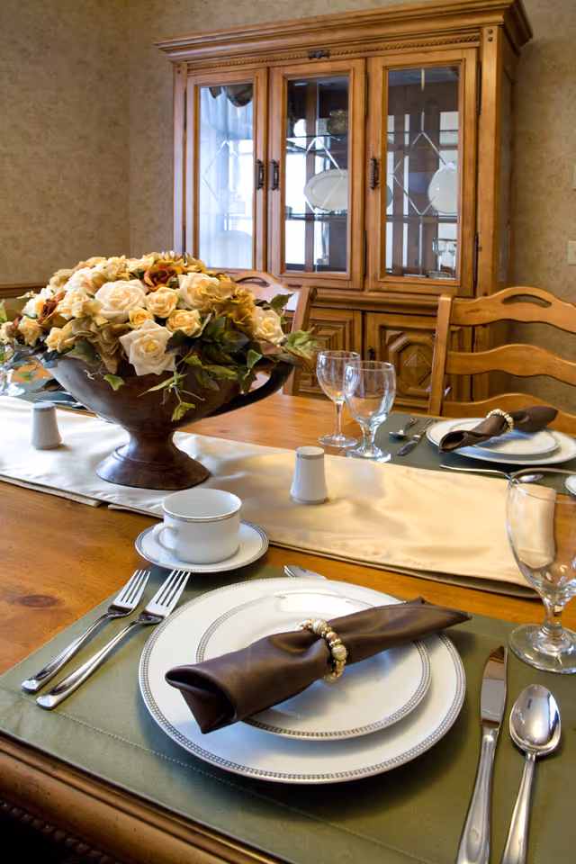 A dining table set for a meal with plates, silverware, glasses, and napkins with decorative rings. A large floral centerpiece with cream and light brown flowers sits in the middle of the table. In the background, there is a wooden china cabinet with glass doors displaying plates and glassware.