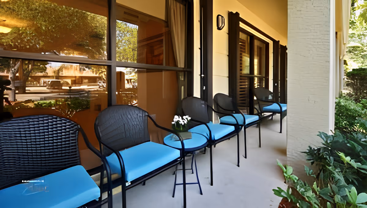 Outdoor patio area with a row of black metal chairs with blue cushions and small round tables, adjacent to a building with large windows and cream-colored walls. Green plants are visible on the right side.