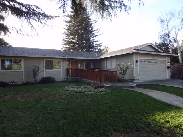 Single-story residential building with beige siding and a gray roof, featuring a wooden ramp leading to the front door, a two-car garage, and a well-maintained green lawn with trees in the background.