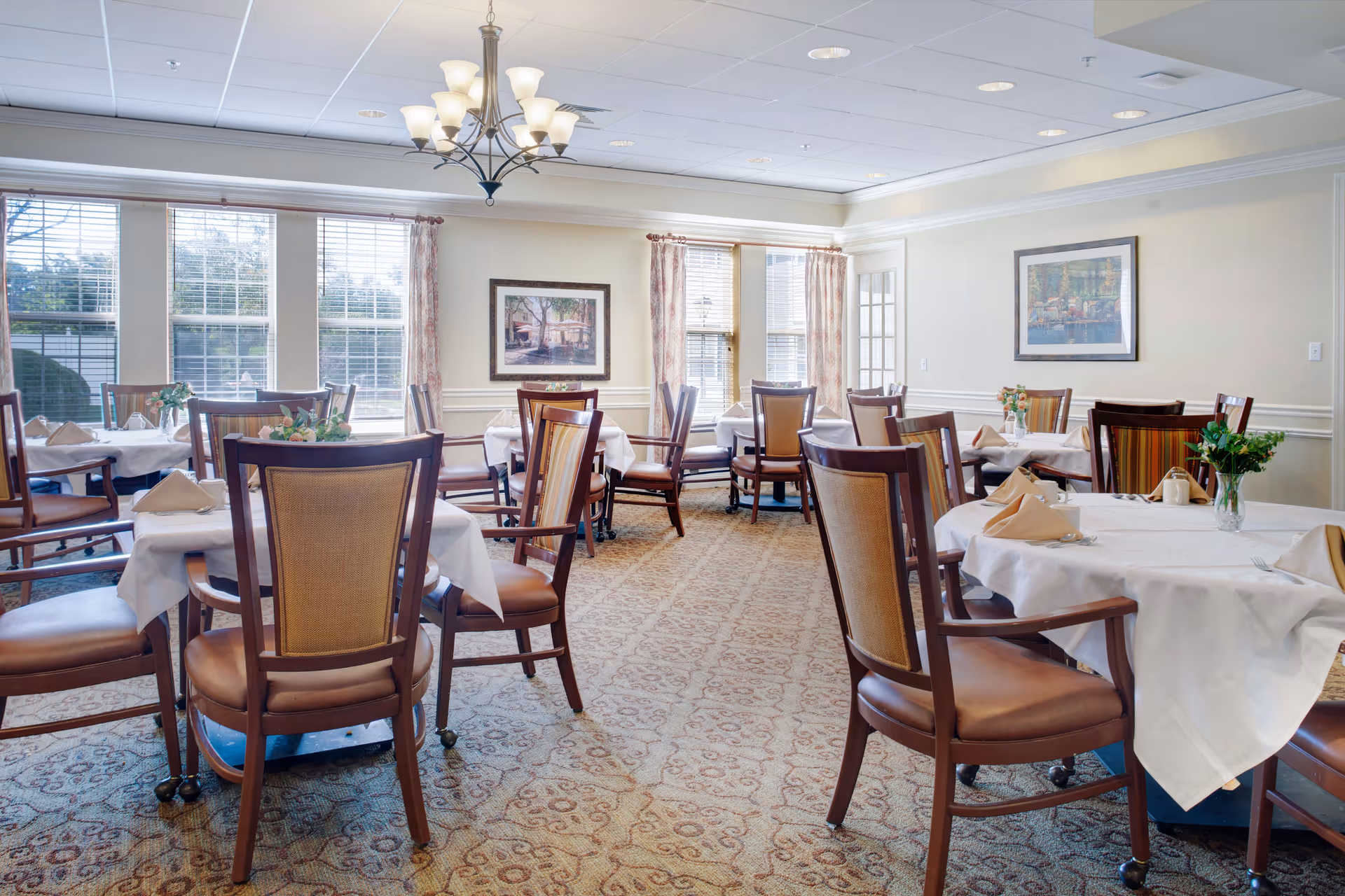 A bright and elegant dining room with multiple round tables covered in white tablecloths, each set with beige napkins and small floral centerpieces. The room features large windows with curtains, framed artwork on the walls, a patterned carpet, and a chandelier hanging from the ceiling.