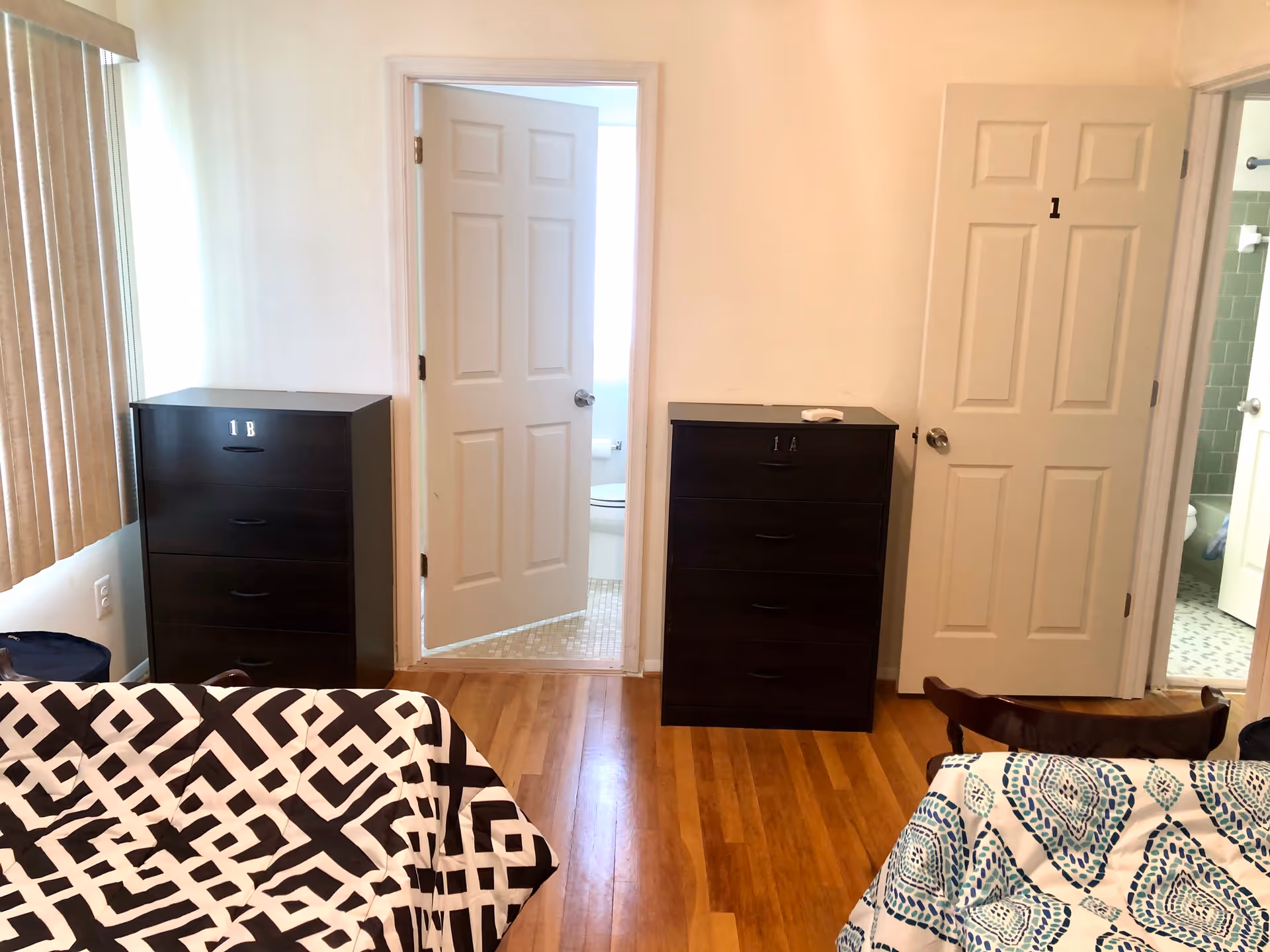 Interior view of a room with two dressers labeled 1A and 1B, two beds with patterned bedspreads, hardwood flooring, and two open doors leading to bathrooms with tiled floors.