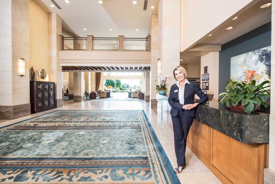 A woman in a dark suit stands smiling behind a reception desk in a spacious, well-lit lobby area with a large patterned rug, plants on the desk, and seating areas visible in the background.