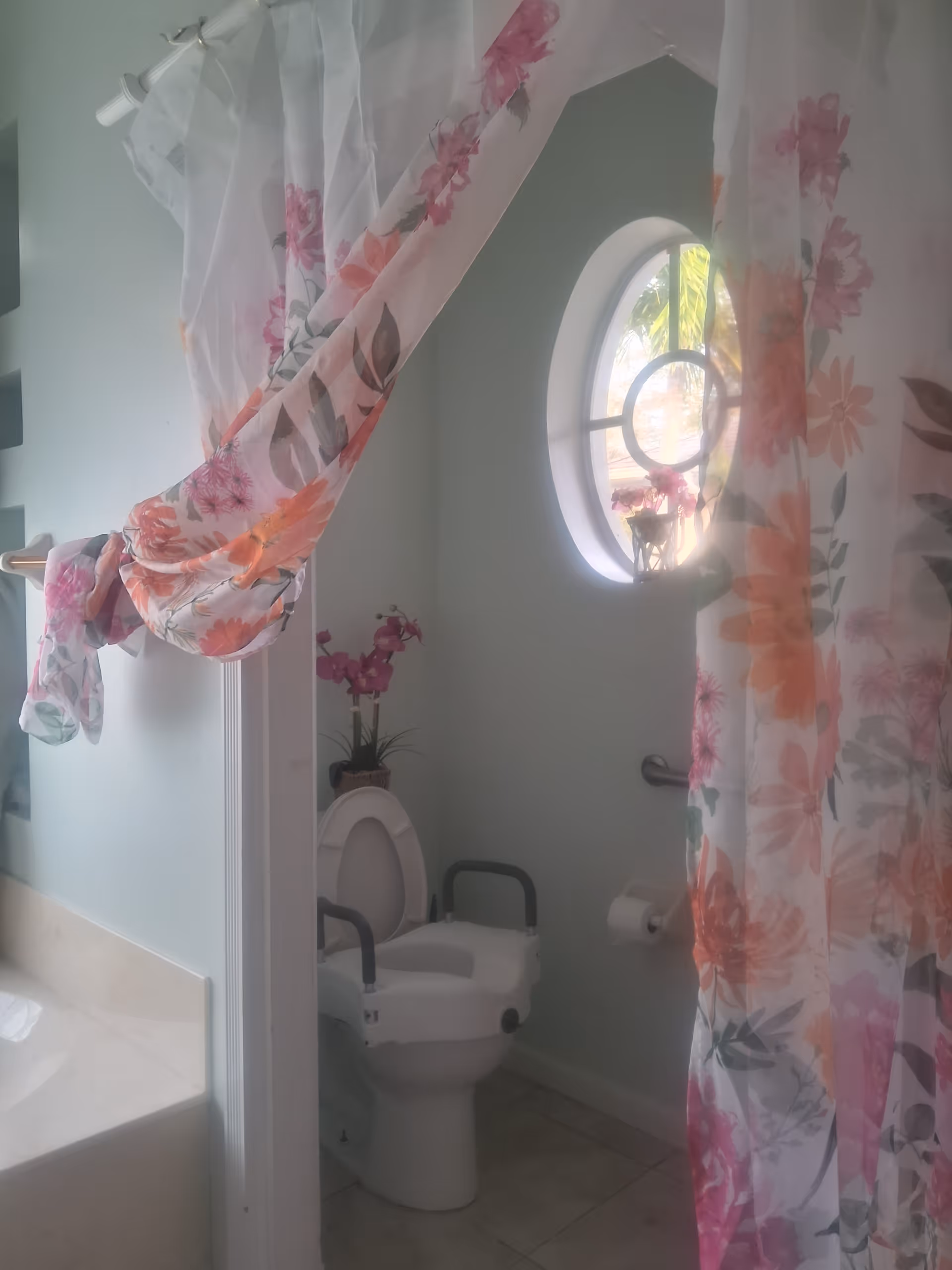 Bathroom interior with a raised toilet seat and armrests, floral shower curtains, and a round window.