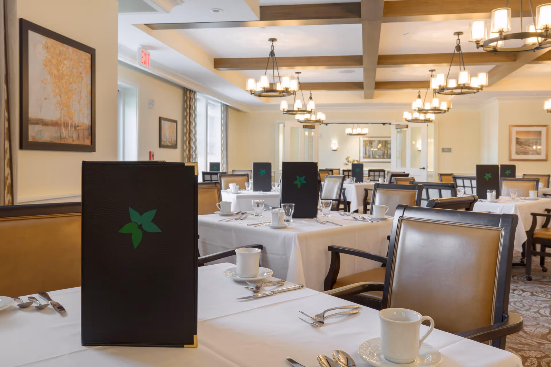 Formal dining room with white-tablecloth tables, place settings, menus bearing a green leaf logo, leather chairs and multiple chandeliers.