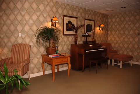 A cozy interior room with patterned wallpaper, a wooden piano with a bench, two framed pictures above the piano, a small wooden side table with books and a plant, a striped armchair on the left, and a cushioned chair with an ottoman on the right.