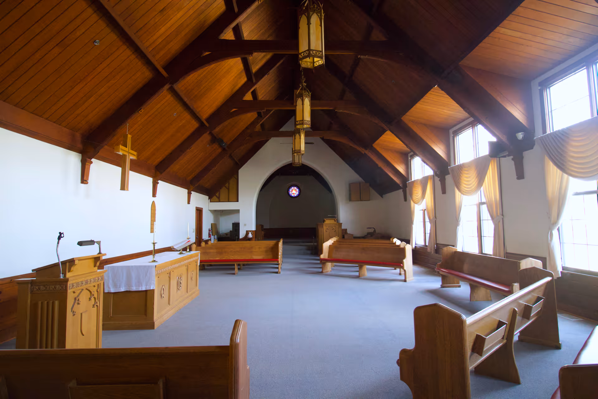 Interior of a chapel with wooden pews arranged facing an altar with a cross on the wall behind it. The ceiling is vaulted with wooden beams and hanging lantern-style lights. Large windows with draped curtains line one side of the room, allowing natural light to fill the space.