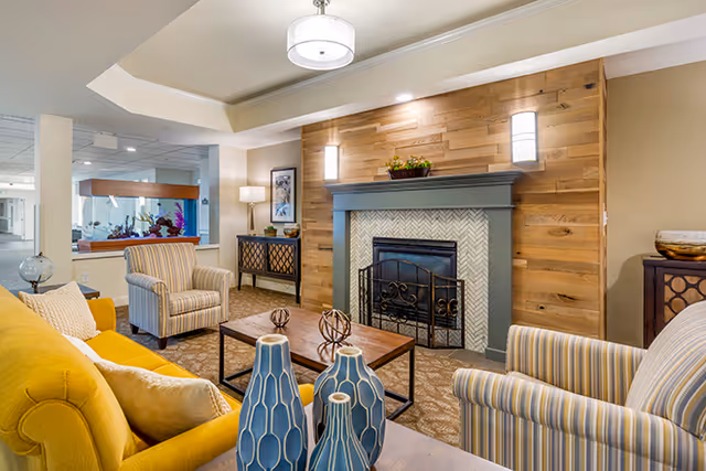 A cozy living room with a yellow sofa, two striped armchairs, a wooden coffee table with decorative blue vases, and a fireplace with a wooden accent wall. The room is softly lit with ceiling lights and a floor lamp, and there is a fish tank visible in the background.