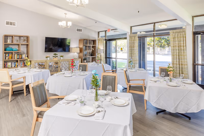 Bright dining room with multiple round tables covered in white tablecloths, each set with plates, glasses, napkins, and silverware. Chairs with wooden frames and gray cushions surround the tables. Large windows with light-colored curtains let in natural light. Shelves with books and decorative items, a wall-mounted TV, and a sideboard with plants and lamps are visible in the background.