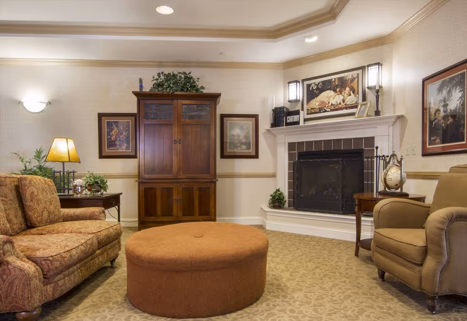A cozy living room with a patterned sofa on the left, a round brown ottoman in the center, and a beige armchair on the right. There is a wooden cabinet against the back wall, flanked by framed artwork. A white fireplace with a black screen is on the right side of the room, decorated with books, a globe, and a framed picture above it. The room has warm lighting and beige patterned carpet.