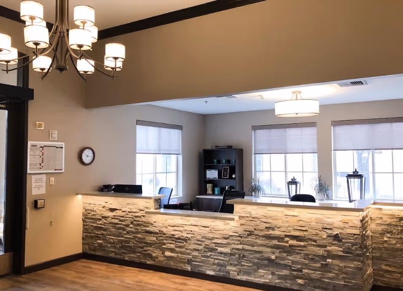 Reception area with a stone-faced counter, two office chairs behind the counter, a bookshelf with decorative items, three large windows with blinds, a wall clock, and a chandelier hanging from the ceiling.