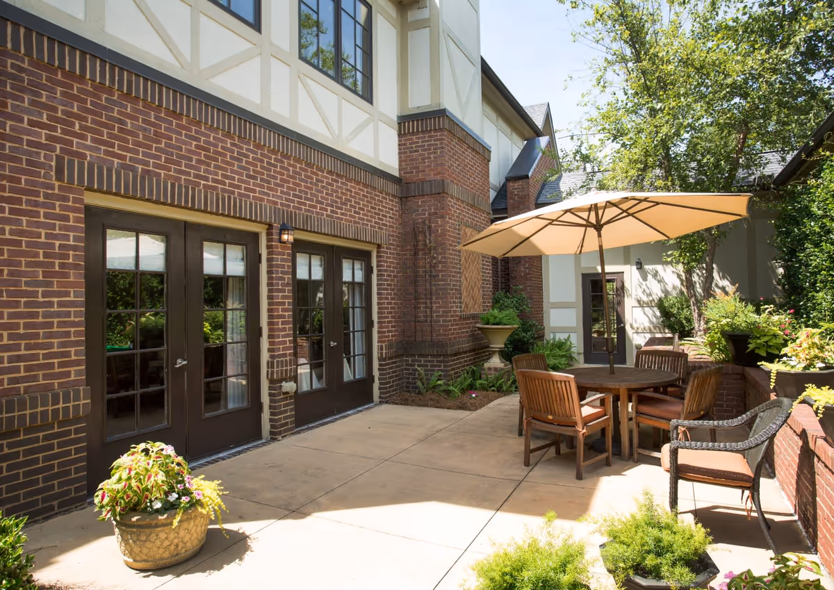 Outdoor patio area at Oak Grove Inn featuring a round wooden table with four cushioned chairs and a large beige umbrella. The patio is surrounded by brick walls and planters with green plants and flowers. The building has a brick and cream exterior with large glass doors and windows. Trees and shrubs provide additional greenery and shade.