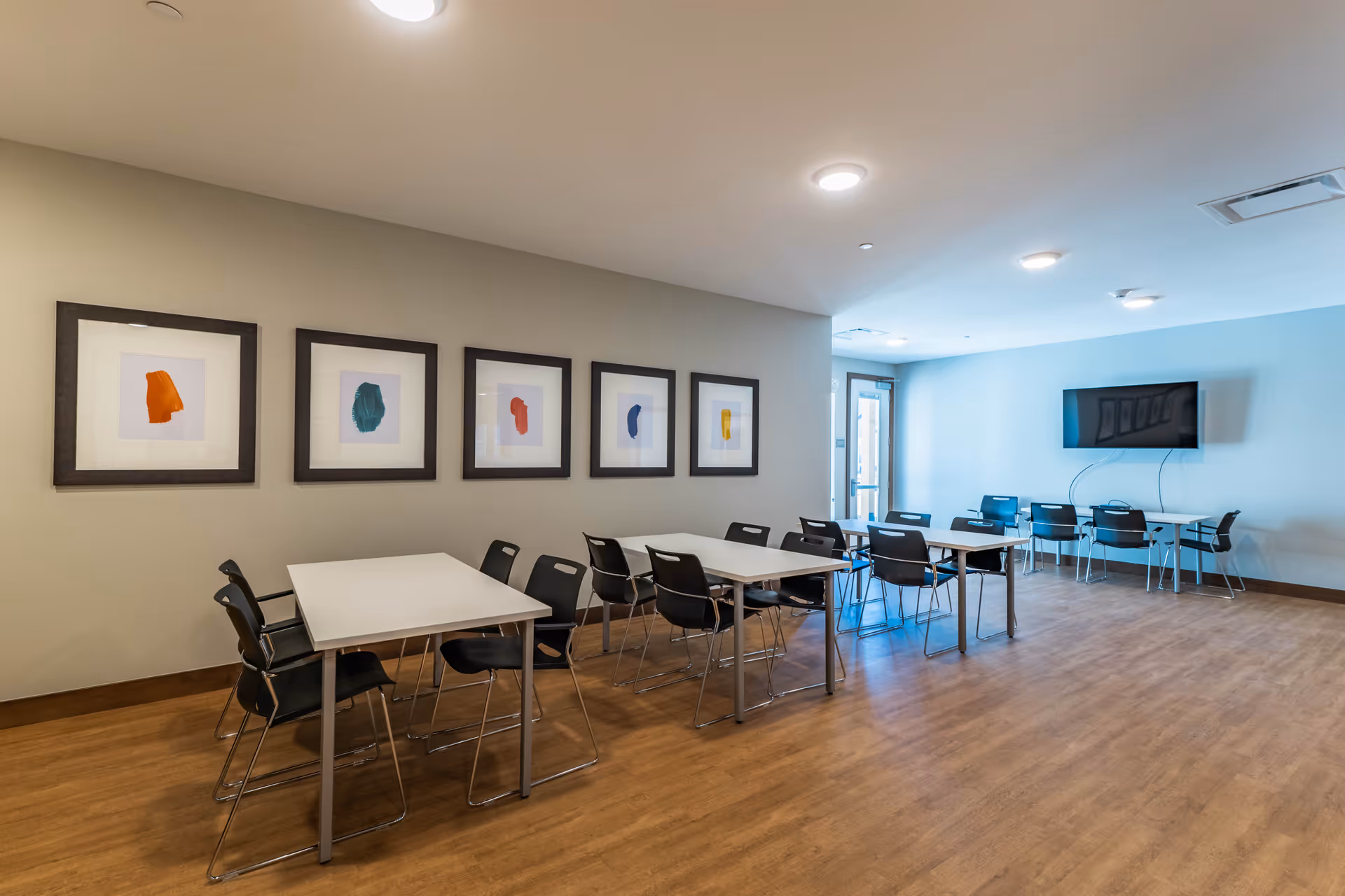 A bright communal dining/activity room with white tables, black chairs, framed artwork on the wall, and a wall-mounted television.