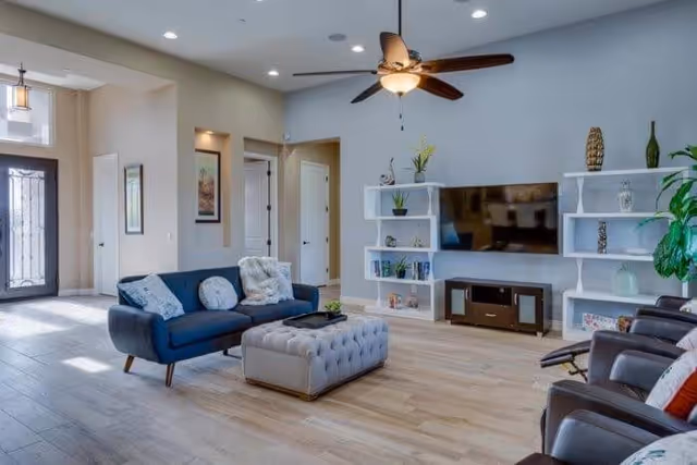 A modern living room with light wood flooring, a blue sofa with cushions, a tufted gray ottoman, and three brown leather chairs. The room features a ceiling fan with lights, a large flat-screen TV mounted on a light gray wall, and two white shelving units with decorative items and plants. There is a large window near the entrance door allowing natural light to fill the space.