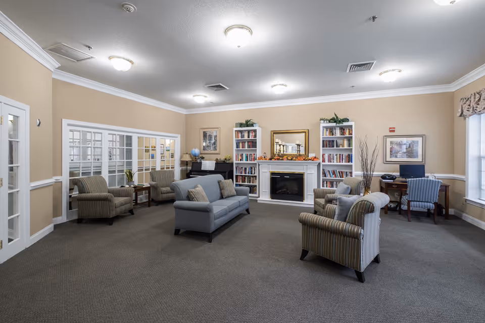 A cozy living room area with beige walls and carpeted floor. The room features a gray sofa and several striped armchairs arranged around a white fireplace with a mirror above it. Two tall bookshelves filled with books flank the fireplace. There is a piano with a globe on it in the corner, and a small desk with a computer and chair near a window with floral curtains. The room is well-lit with ceiling lights and has white crown molding and chair rails.