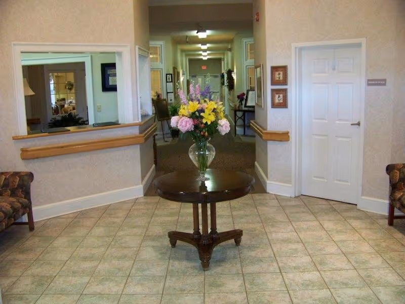 Interior view of a hallway in an assisted living facility with a round wooden table in the foreground holding a glass vase filled with colorful flowers. The hallway extends into the background with doors and framed pictures on the walls. Two patterned armchairs are partially visible on either side of the image.