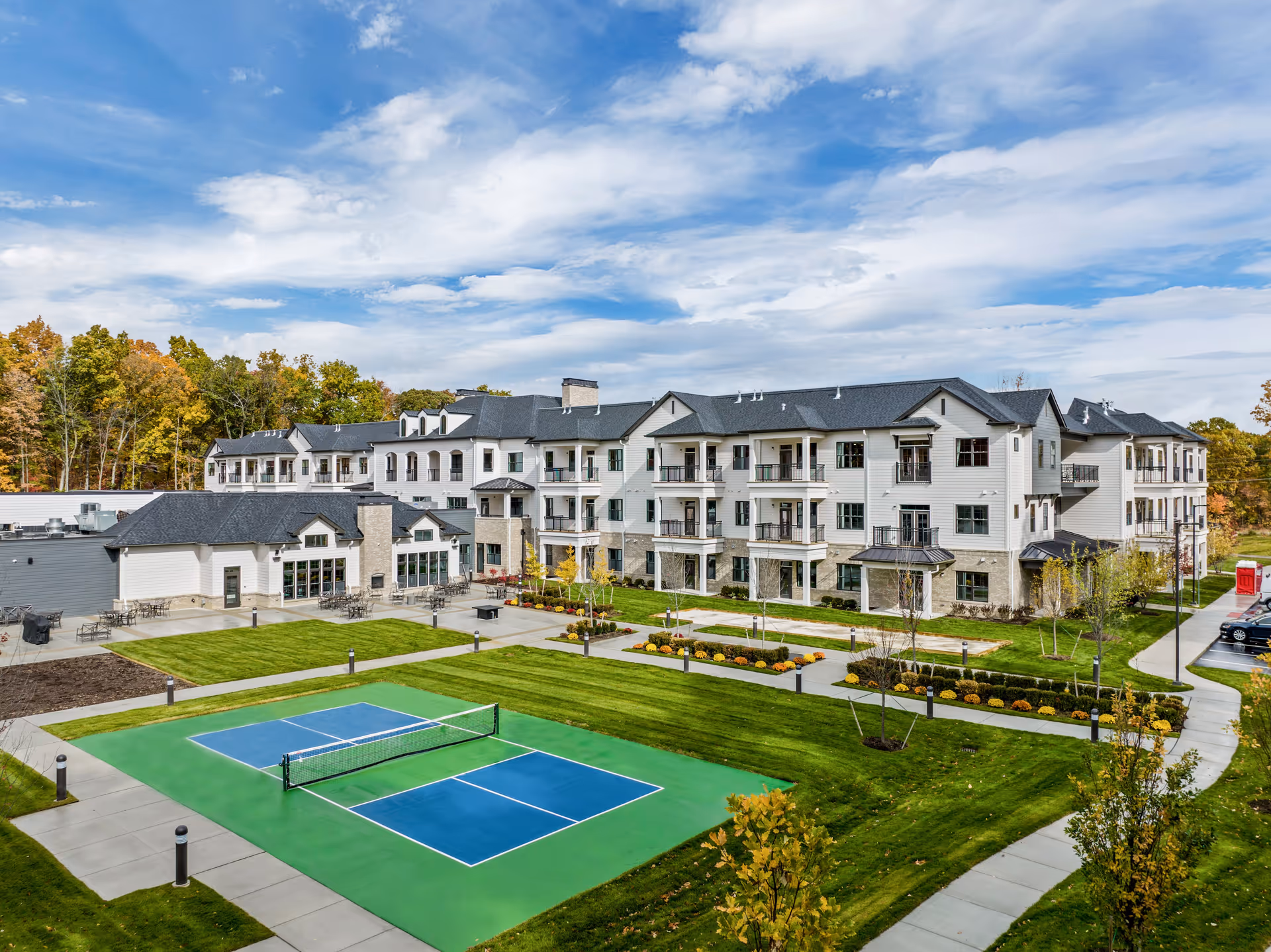 Exterior view of Monark Grove Clarkston, showing a large three-story building with balconies, surrounded by well-maintained green lawns and landscaped gardens. In the foreground, there is a blue and green pickleball court with a net. The sky is partly cloudy with patches of blue.
