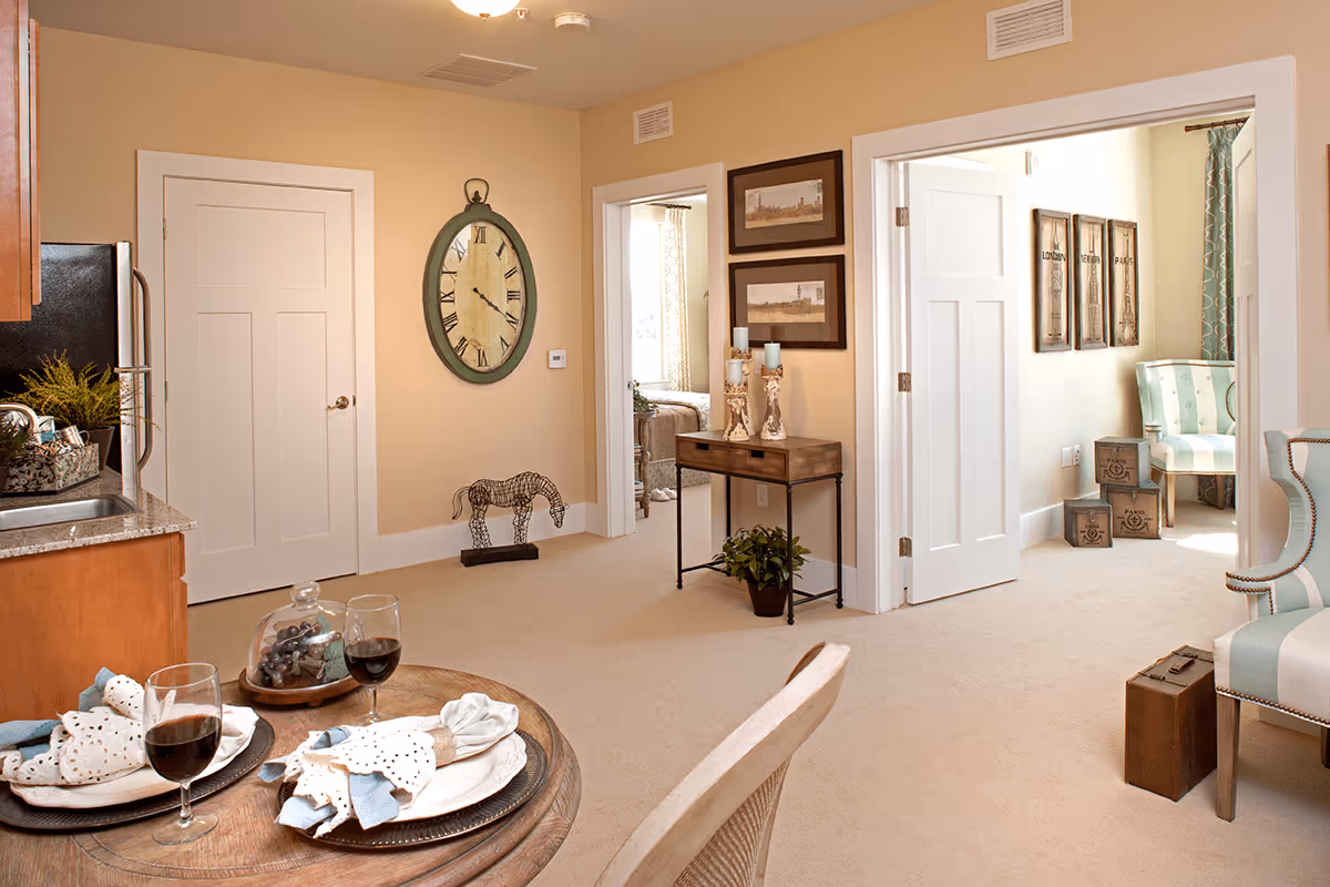 Interior view of a cozy assisted living facility showing a small dining table set for two with wine glasses and napkins, a kitchen area with a sink and cabinets on the left, a beige carpeted floor, a large wall clock, decorative horse sculpture, a small console table with candles and framed pictures on the wall, and two open doorways leading to a bedroom and a sitting area with a striped armchair and decorative boxes.