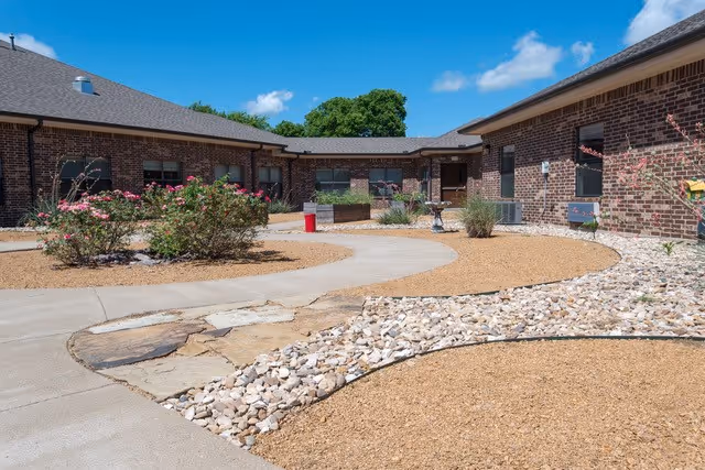 Outdoor courtyard area at Kemp Care Center with a curved concrete walkway, landscaped with rocks, mulch, and flowering bushes. The courtyard is surrounded by single-story brick buildings under a blue sky with some clouds.