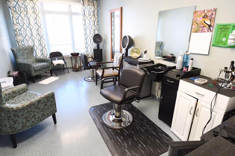 A bright room with large windows covered by patterned curtains, featuring salon chairs, a hair washing station, mirrors, and various hair care products, along with comfortable armchairs and a small table in the corner.