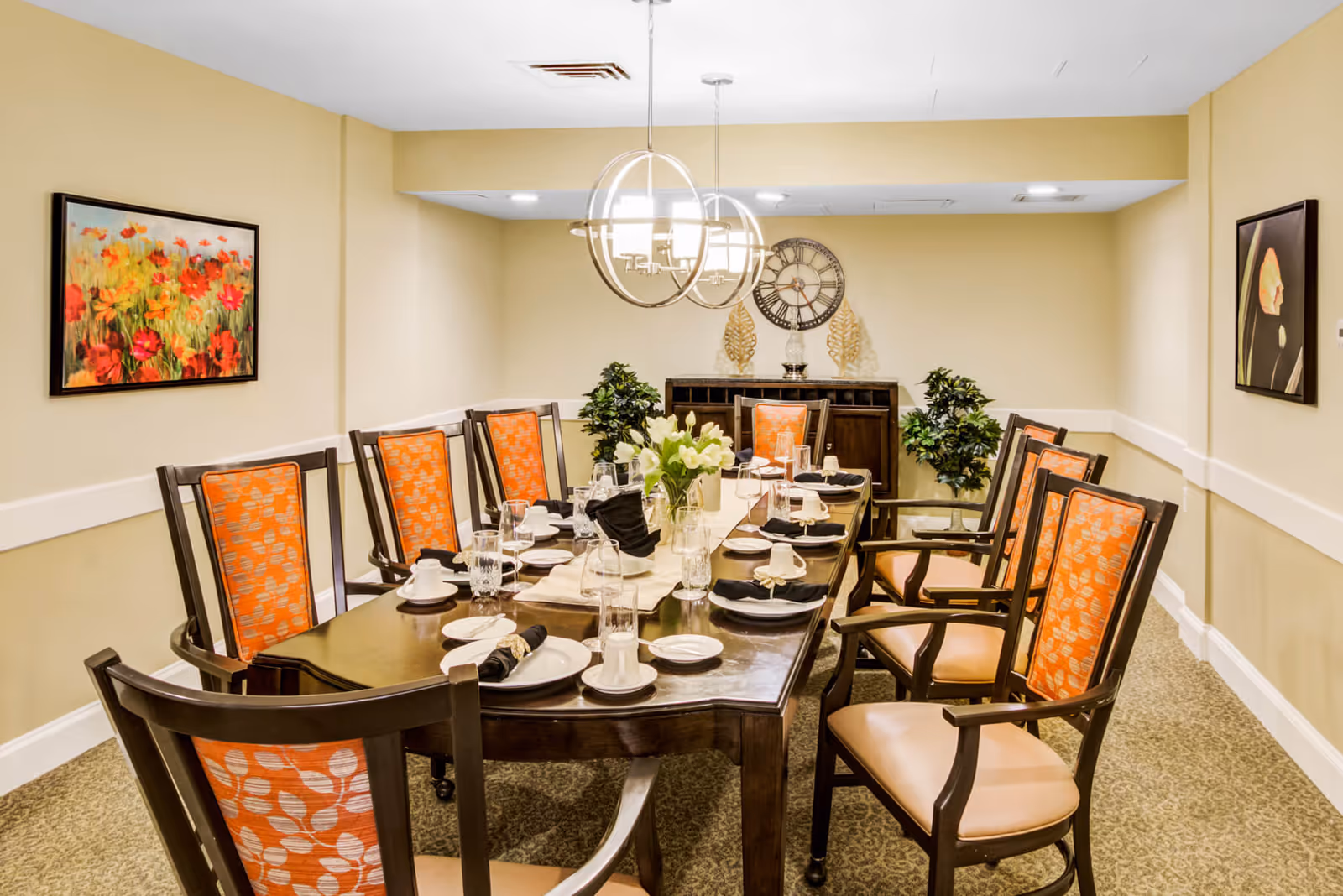 A formal dining room with a long dark wooden table set for a meal with plates, glasses, napkins, and a centerpiece of white flowers. The room has beige walls, two modern circular chandeliers hanging from the ceiling, and orange cushioned chairs with wooden frames arranged around the table. There are two framed paintings on the walls and two green plants near a sideboard with decorative items including a large clock.