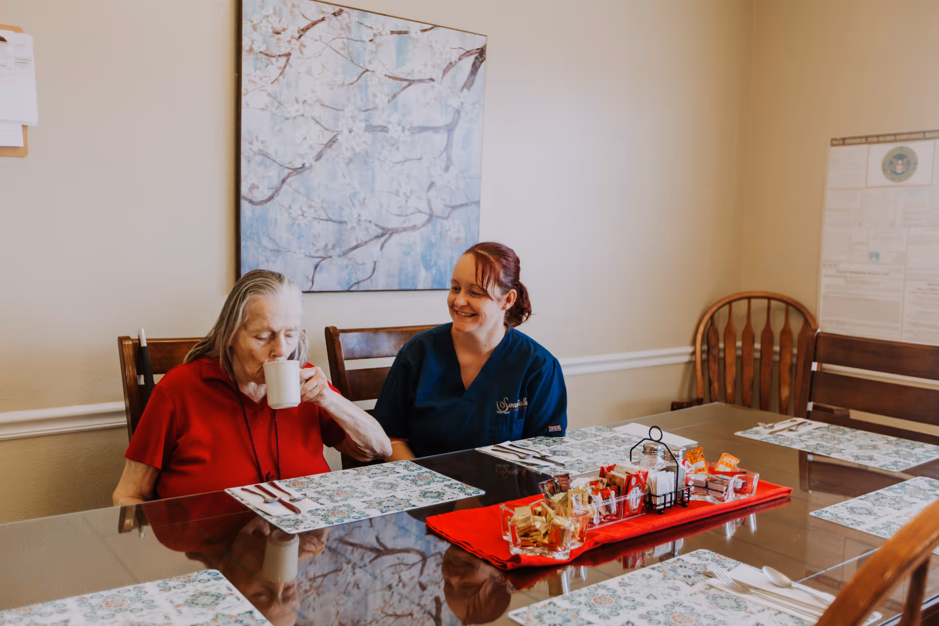 An elderly woman in a red shirt drinks from a white mug while sitting at a dining table with a caregiver in blue scrubs smiling beside her. The table is set with placemats, utensils, and a red tray holding various condiments and packets. The room has beige walls with a floral painting and a bulletin board in the background.