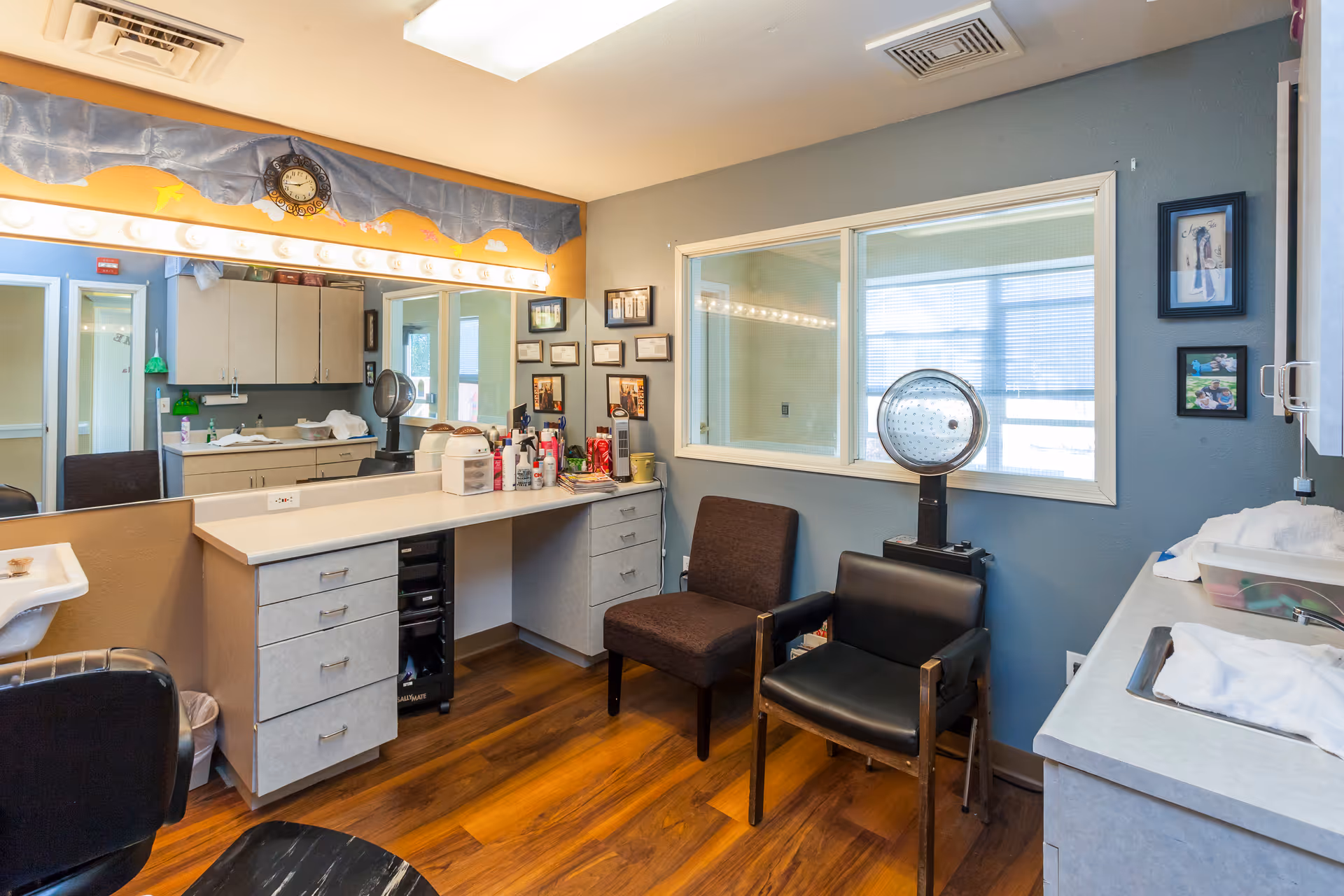 Interior view of a senior living facility's grooming or beauty room with a large mirror surrounded by lights, a countertop with various hair and beauty products, two chairs, a hair dryer, and framed pictures on the walls. The room has wood flooring and a window looking into another room.
