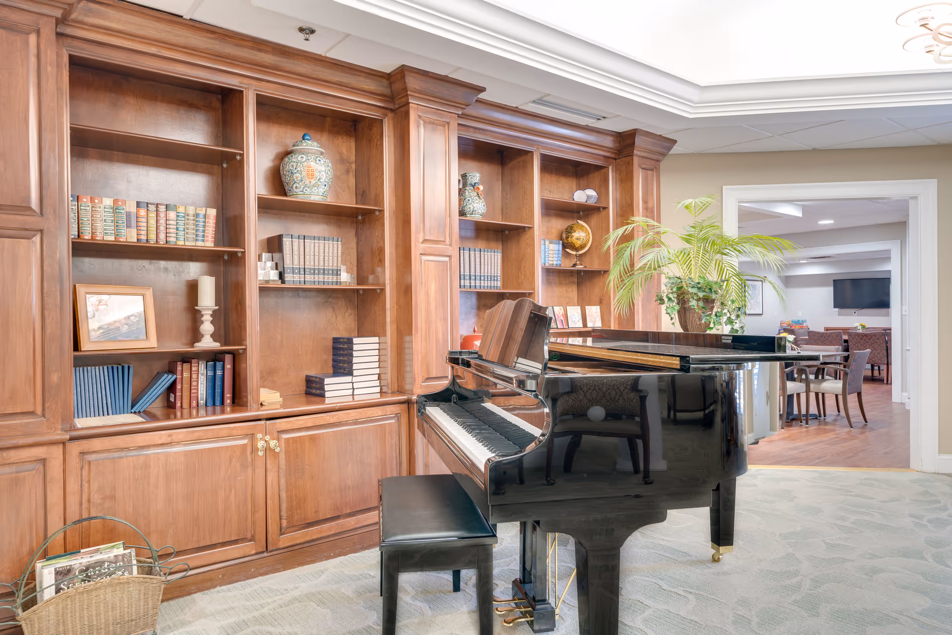 Common area with a black grand piano in front of wood bookshelves and a seating/dining area visible through a doorway.