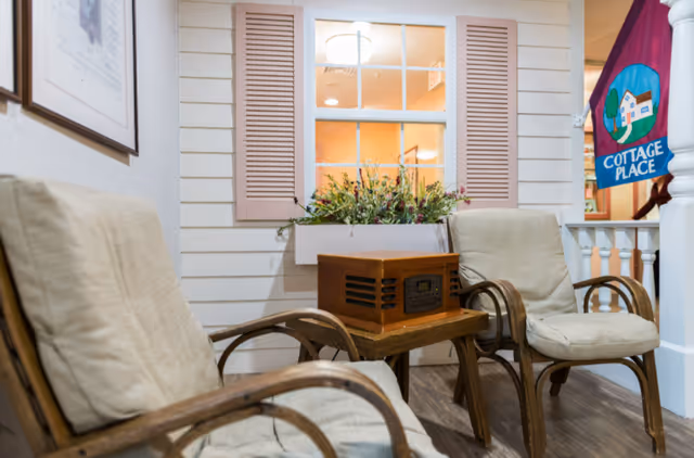 Two cushioned wooden armchairs with a small wooden table between them holding a vintage-style radio, set against a wall with a window box of flowers and pink shutters. A colorful Cottage Place flag is visible in the background.